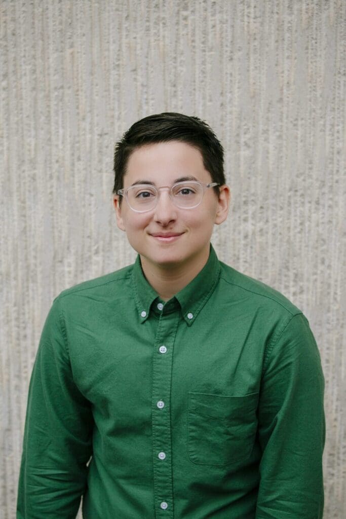 A short black haired person in glasses and green button up shirt smiling.