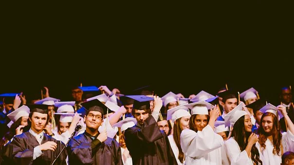 This image looks like one from a graduation ceremony, there are a lot of girls and boys. The boys are wearing black gowns, while the girls are wearing a white gowns. They all touch the graduation hat that they are wearing.