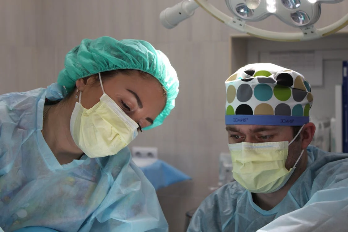 This image is from an operation theatre and one can see two doctors wearing surgical masks, head caps, and scrub suits appear to be looking at the operation table.