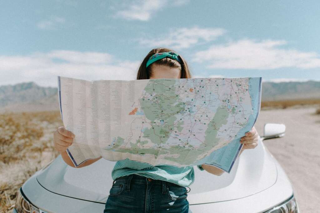 This image is in the daytime, with a bright sunny day and clear sky showing a young girl looking at a map while sitting on the bonnet of a white color car. While she is holding the map in front of her, only her forehead is visible.