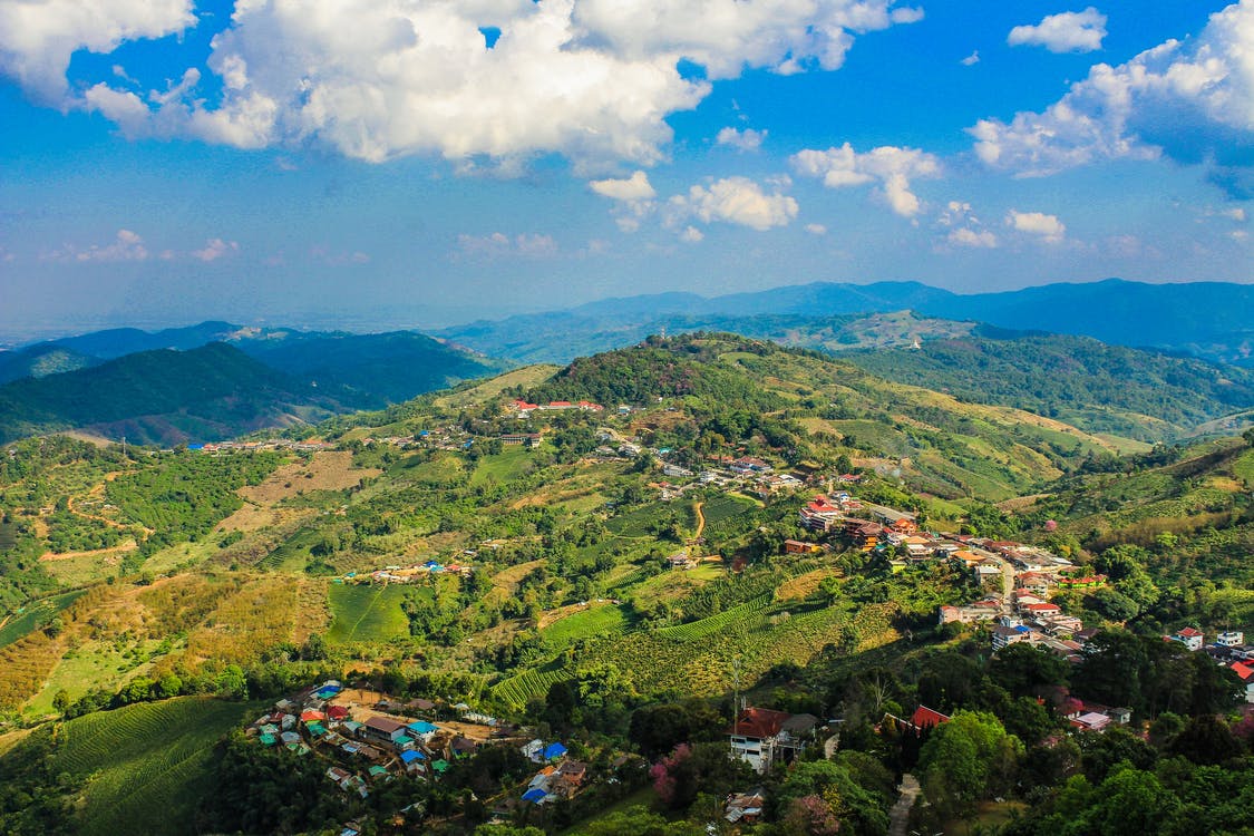This image shows a picture taken from the top of a mountain, below there is greenery with houses. We can also see blue sky and clouds.