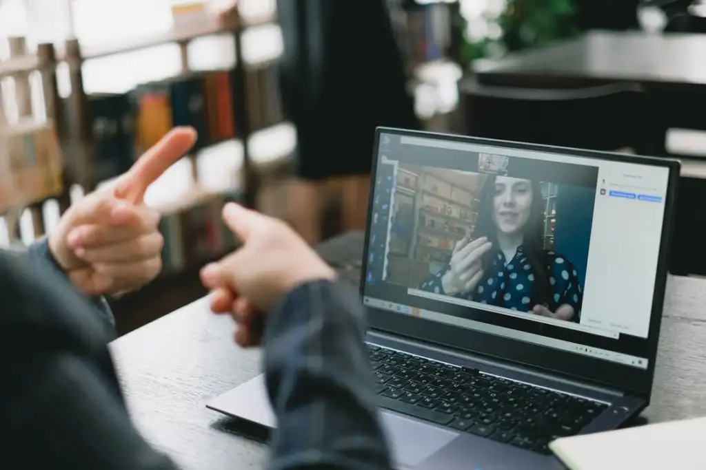 This image shows a video call happening between a lady on the laptop screen sitting in front of the laptop. They are communicating through sign language.