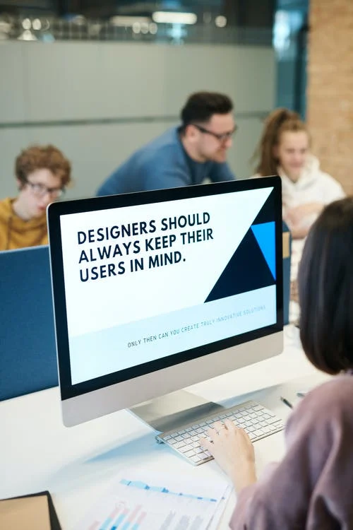 This image appears to be an office setup. A woman is working on her computer and the screen, and it's written as " Designers should always keep their users in Mind". On the opposite side to the woman, there are three more people working, two women and one man.