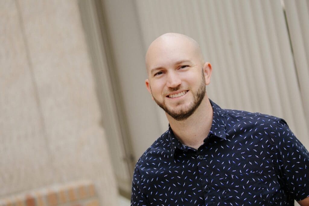 A bald haired person in a blue buttoned up shirt and white polka dots smiling.