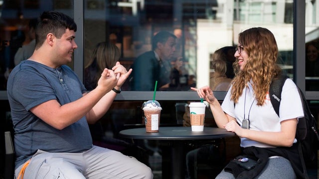 This image shows two people, a man and a woman sitting outside a restaurant with two coffee on the table. They are a sign language conversation.