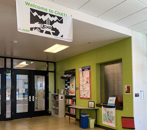 This image is of a hallway with a green wall and an adjacent glass wall with a glass door. Against the green wall, there is a table, a bookshelf, and some posters. At the entry of the hallway, there is a banner hanging with the text " Welcome to Chat" and a black and white illustration of two hands, a bowl with steam coming from it & cat approaching the bowl.