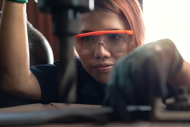This image is of a woman wearing safety glasses looking & trying to drill on something which appears to be a metal rod.