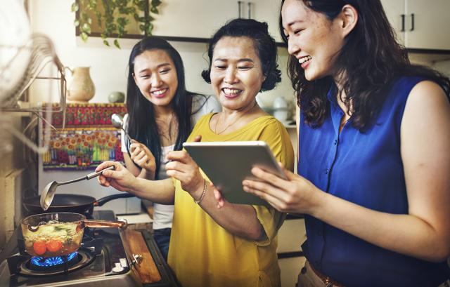 This image shows a mother with her two daughters in the kitchen. The mother is cooking, while one of the daughters is showing something on the iPad and everyone is laughing about it. The family seems of Asian origin.