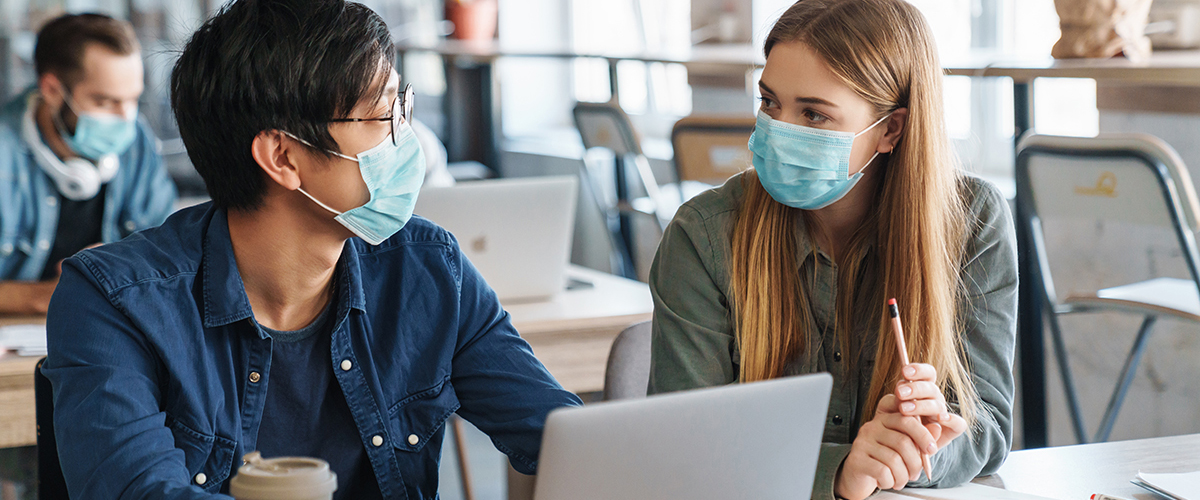 This image shows two students wearing surgical masks who appear to be talking. They have a laptop in front of them and a few notebooks to make a note while studying.