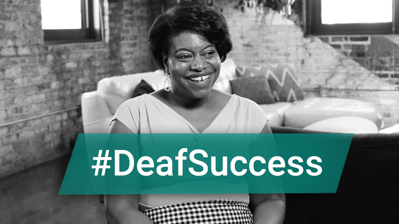 This is a black and white image of a woman sitting on a chair in a room with sofas in the background, She has short hair and a big smile on her face. On the Image, there is a hovering text " #DeafSuccess"