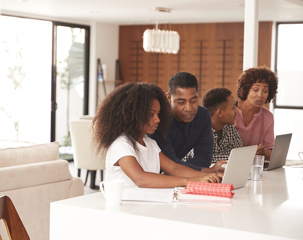 This is an image of a family of four in their house. It appears that the dad is helping the daughter with something on her laptop, while the mom is helping the son with something that is on his laptop. Both the Mom and Daughter have long curly hair.