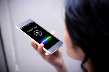 This is an image of a woman trying to use the voice-activated search or command on a mobile phone. The mobile phone display shows a " Speak Now" with a Mic icon.