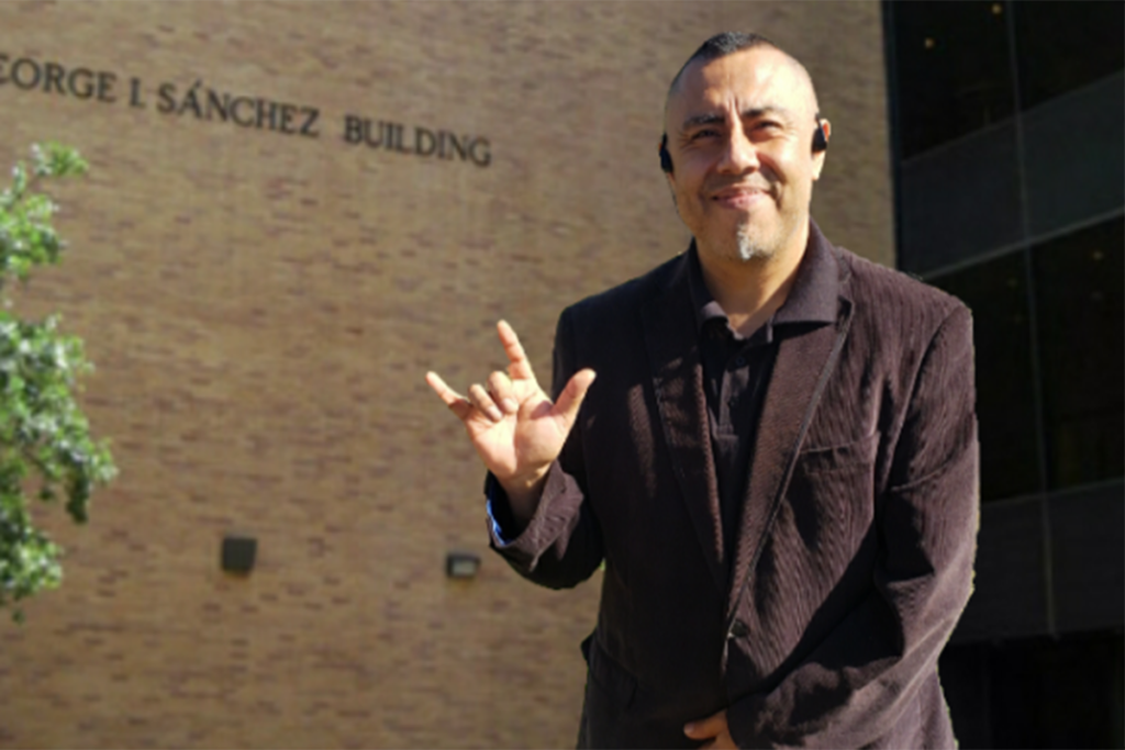 This image is of a man named Crescenciano Garcia who is standing in front of a building on which the name " George L Sanchez Building " is mentioned. He appears to be wearing a brown jacket with a T-shirt inside and is sporting an "I Love You" sign with the fingers on his right hand.