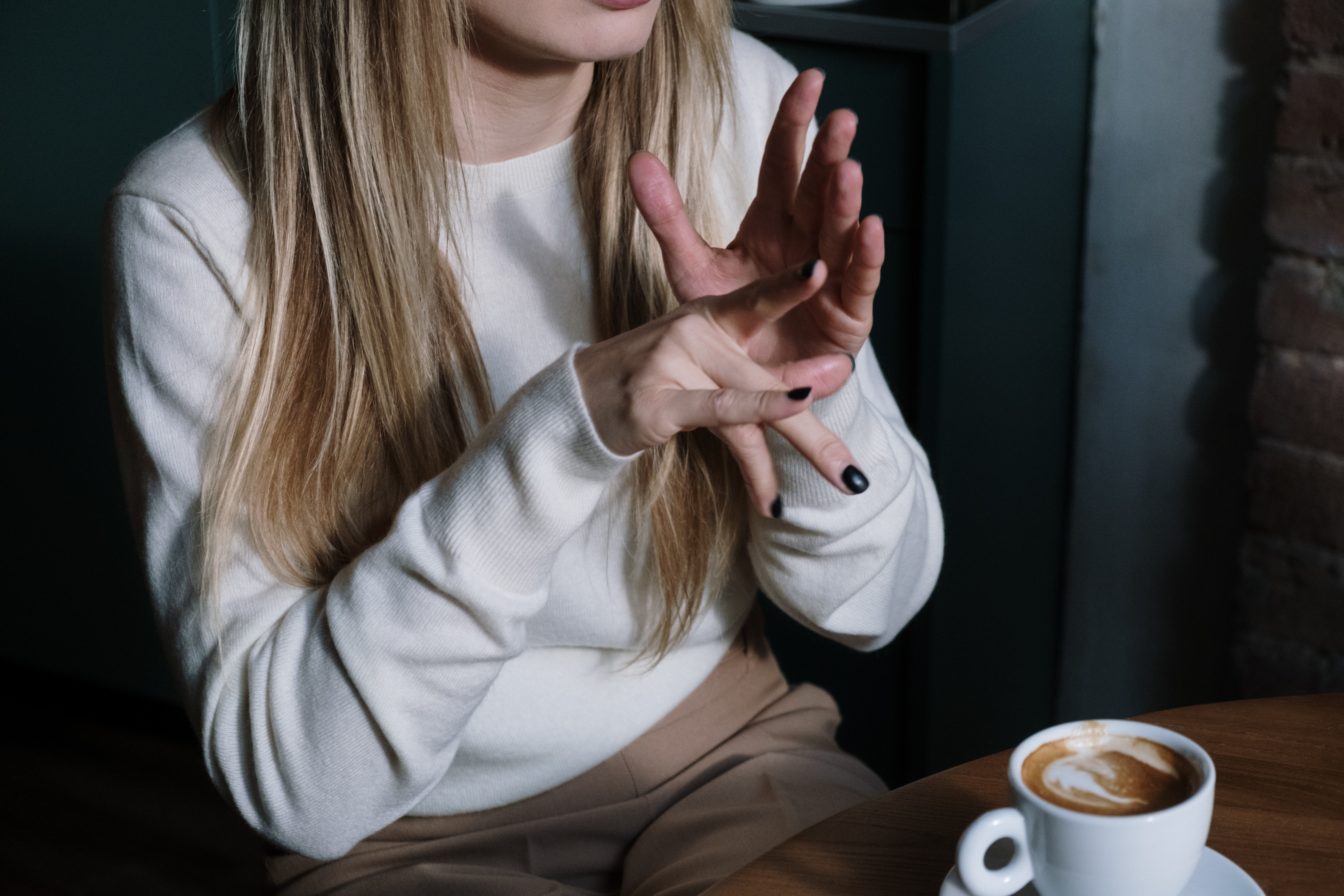 This image shows a woman dressed in a white long sleeve t-shirt sitting in a coffee shop with coffee on the table. She appears to be showing or gesturing with her hands to someone.