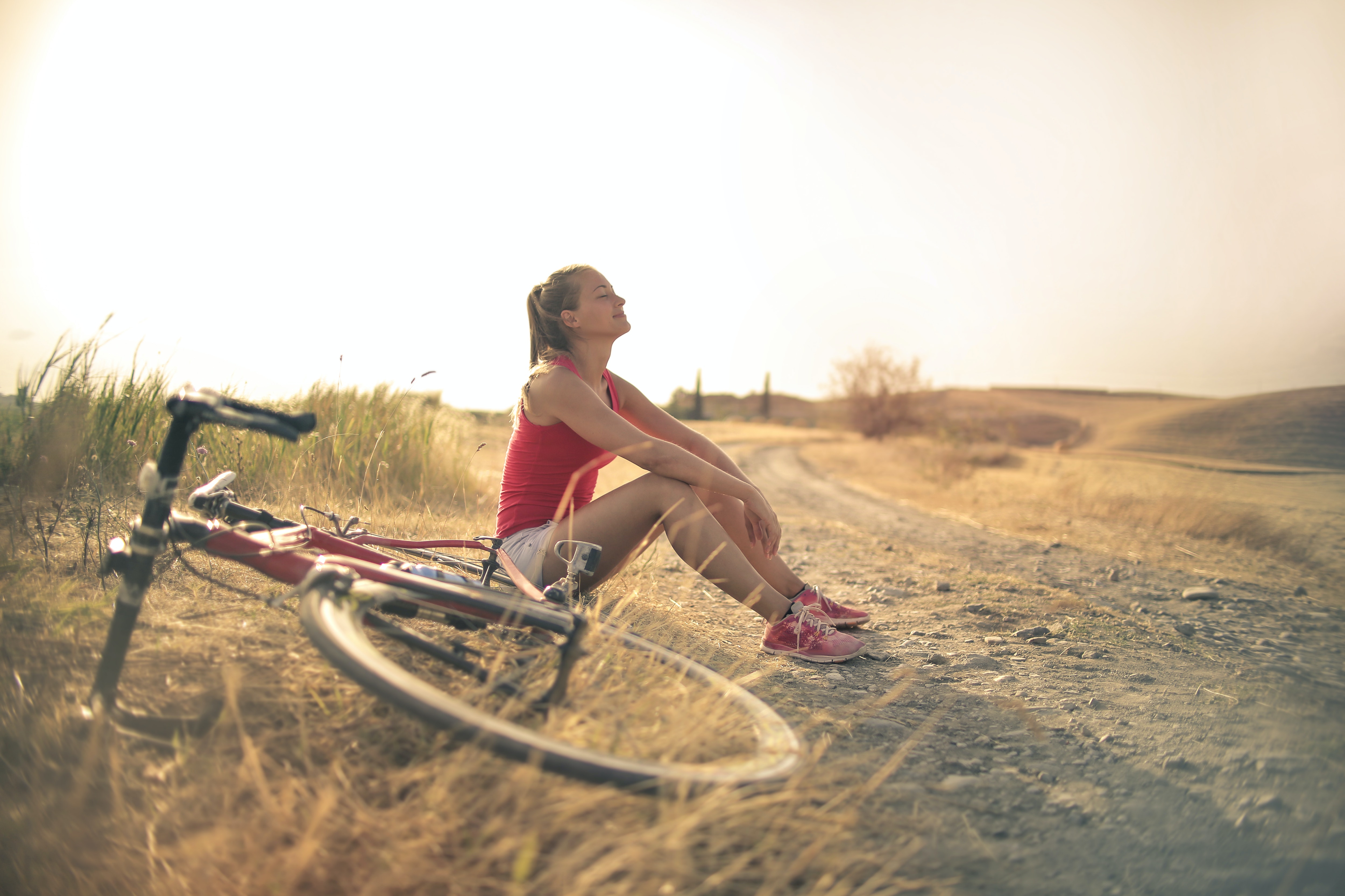 This image is taken in the daytime with the sun shining bright. Women are sitting on the ground after a long bicycle ride. She is wearing a red sports top and shorts, while her bicycle is on the ground next to where she is sitting.