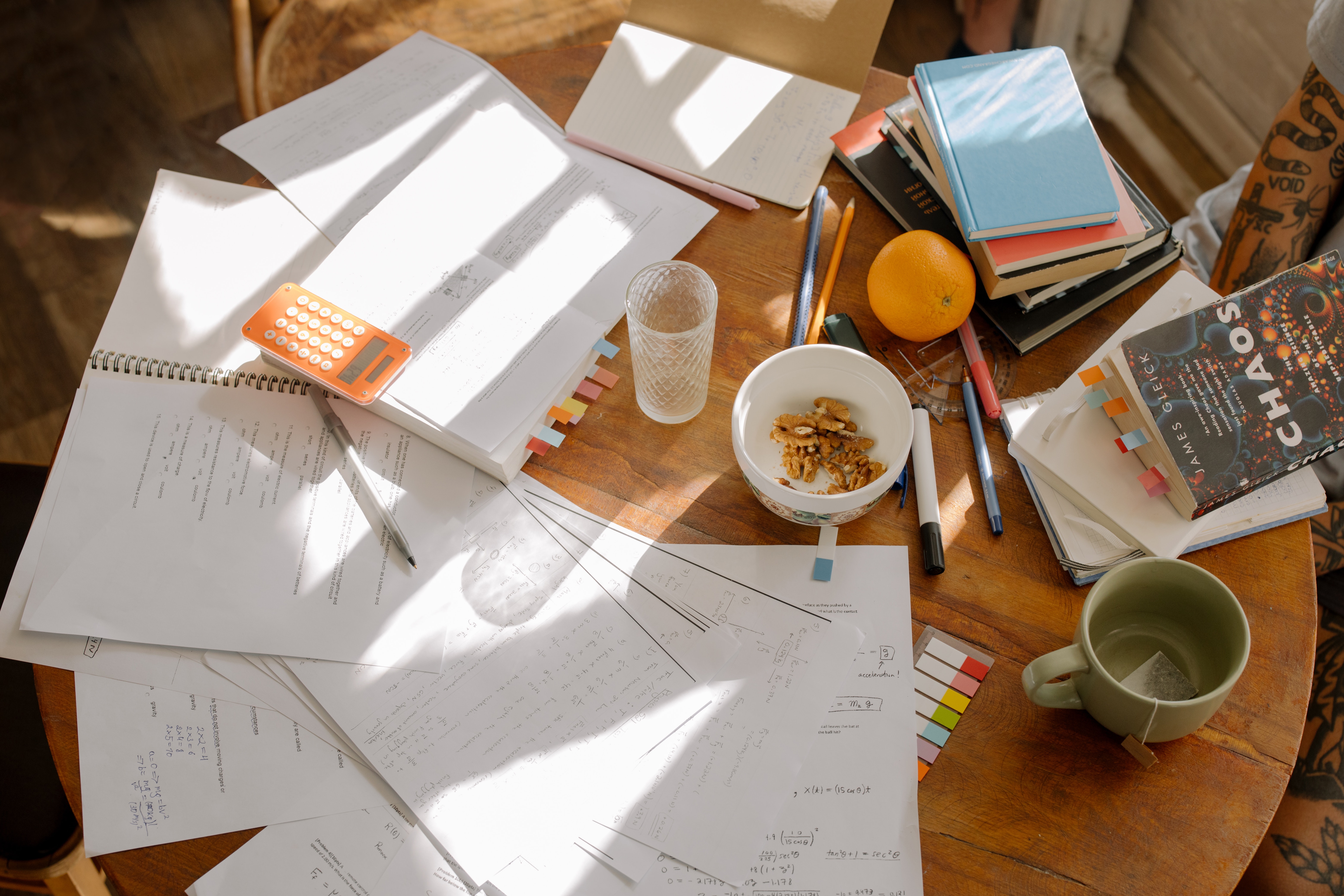 This image is taken from the top and has a round table with papers, books, pens, pencils, a calculator, one empty green cup, an empty glass, one orange, and a bowl of some leftover walnuts.