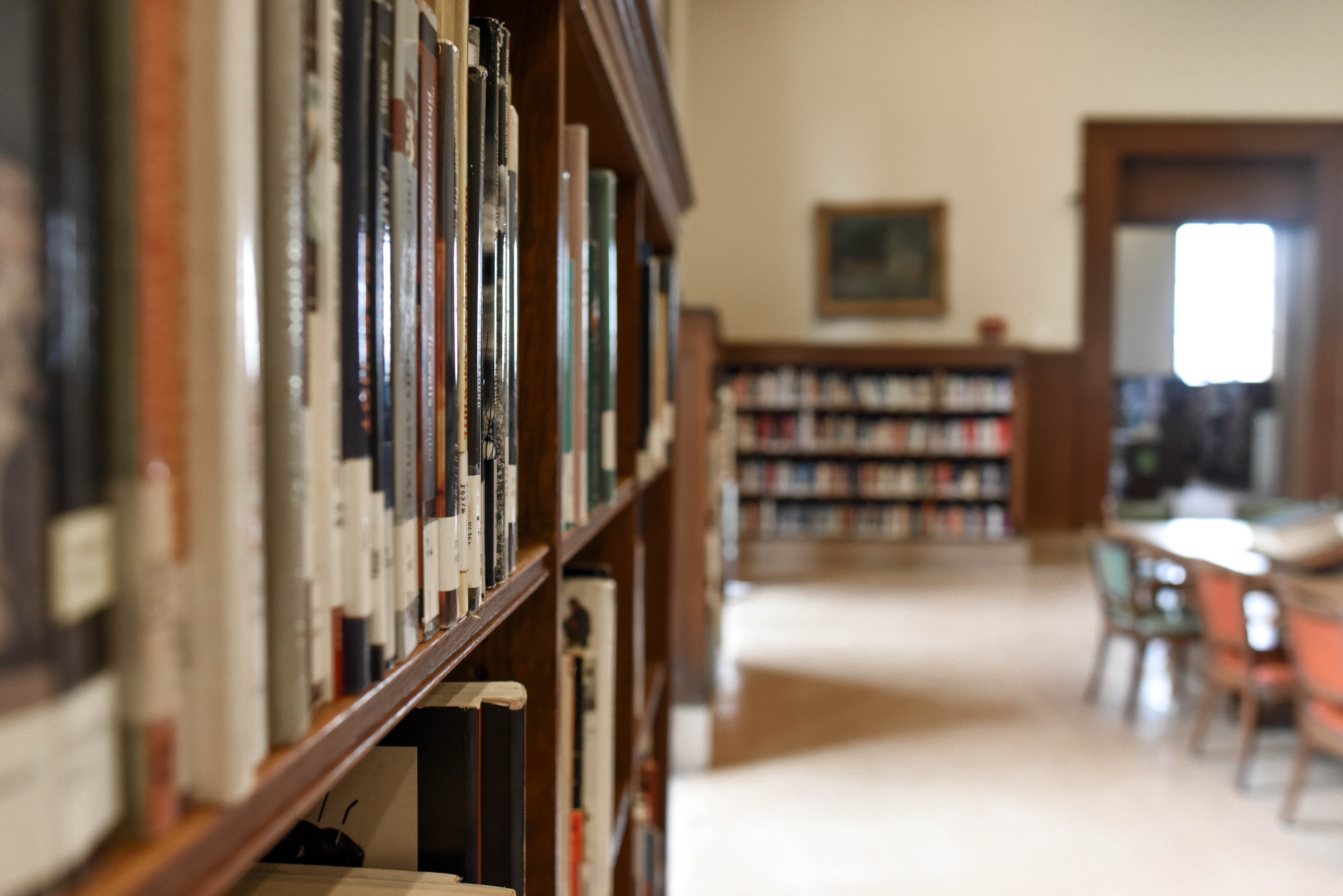 This is an image taken in a library. In the background, there is the blurred image of chairs and bookshelves, while the focus of the image is on a bookshelf with books on the left side.