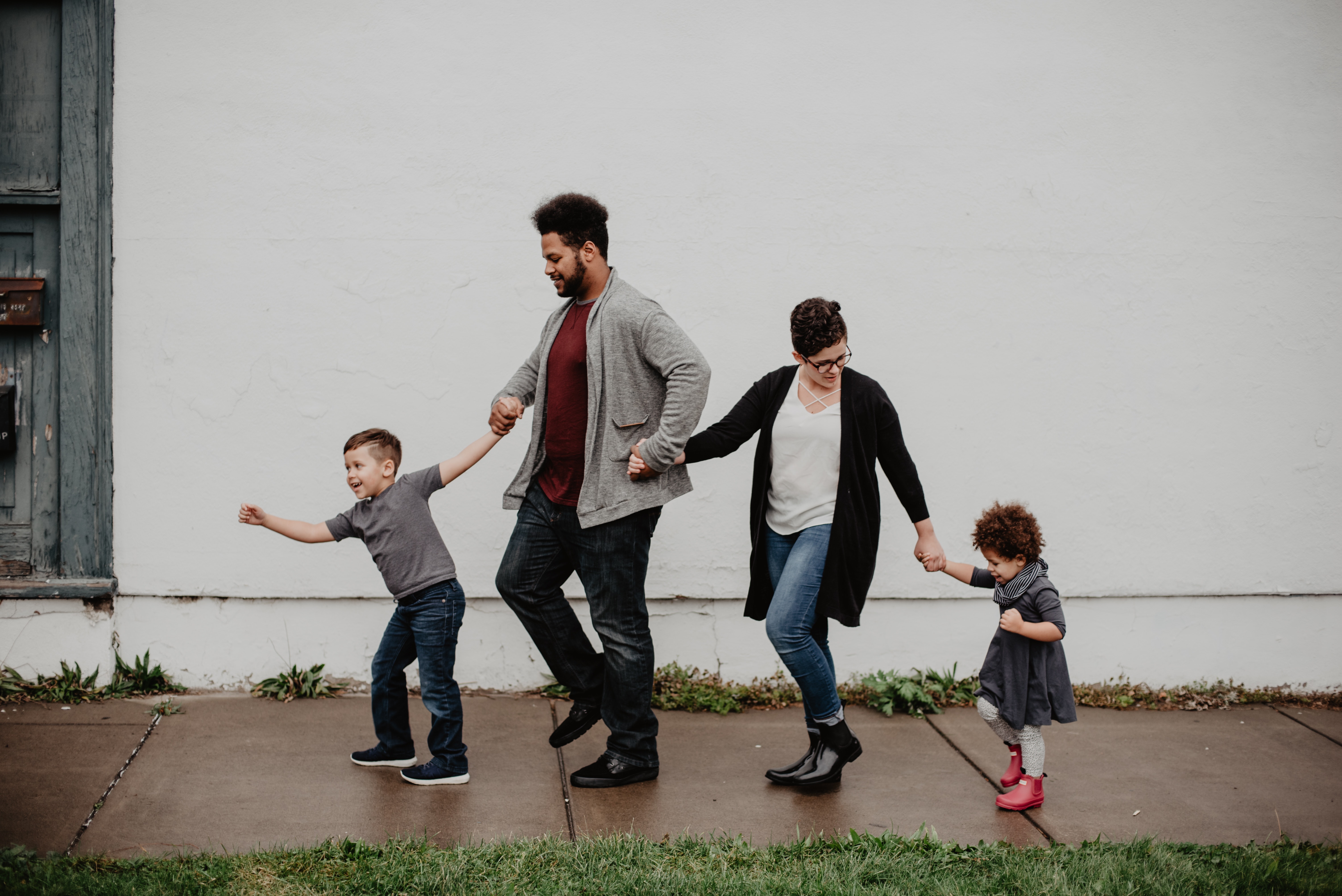 This image is of a family of four walking on the pavement. All four of them are walking holding hands. There are two kids, one boy, and one girl child. The pavement appears to be wet, which may be from the earlier rain. Both parents are wearing sweatshirts on top of the t-shirts.