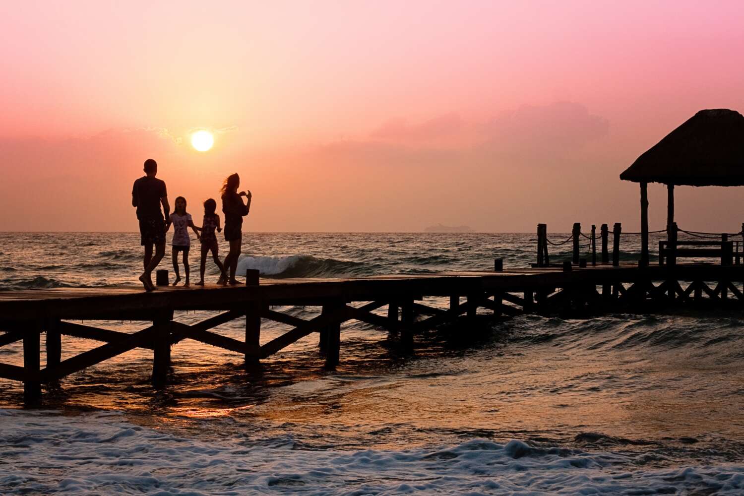 This image is taken around the time when the sun is about to set. It shows a wooden walking area near a beach and there is a family of four walking on it.