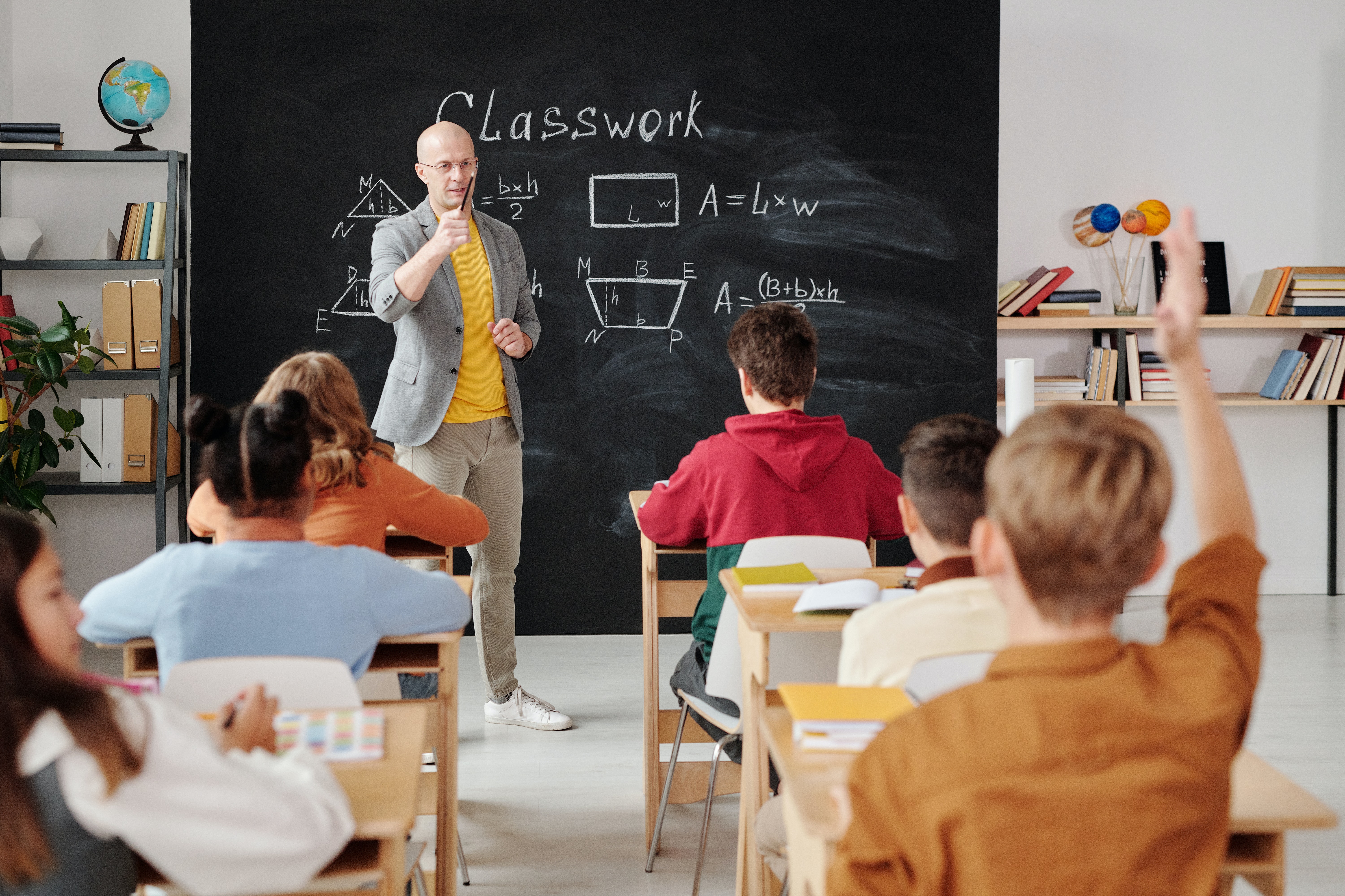 This image shows a teacher in a classroom. He is wearing a grey jacket, a yellow t-shirt, jeans, and white shoes. He is supposedly asking something from the students and one of the students has raised his hands. Behind the teacher, there is a blackboard that mentions " Classwork" and some formulas.
