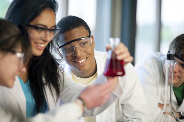 This image shows four people wearing lab coats and safety goggles. There are two men and two women. One of the women is holding a beaker with a red liquid in it, while others are staring at it.