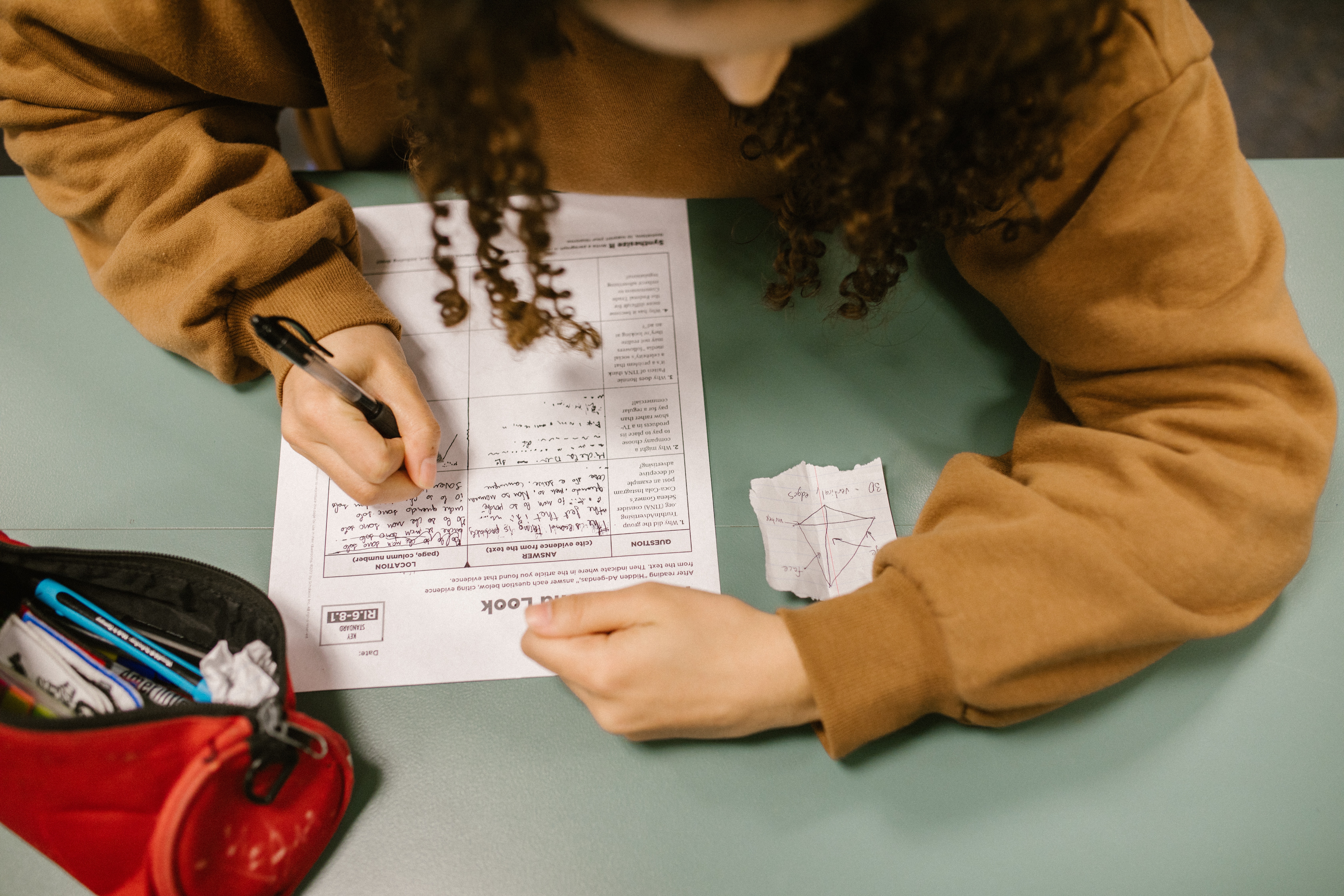 This image shows a young girl writing a test paper. She has a small piece of paper kept next to the test paper. It appears that she is trying to replicate the image on the piece of paper in a section of the test paper.