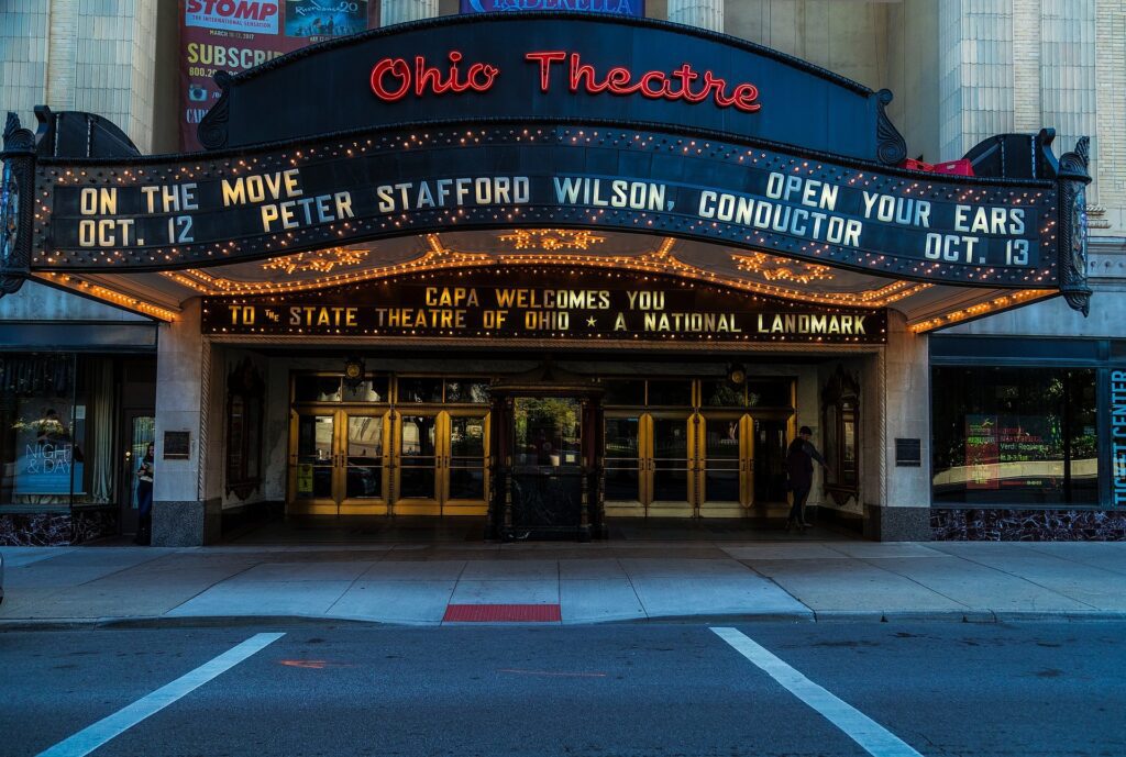 This is a rectangular image. This image is of the entrance of "The Ohio Theatre". Below the theatre name there is the text " ON THE MOVE... OCT 12, PETER STAFFORD WILSON, CONDUCTOR . OCT.13". Below that there is the text " CAPA WELCOMES YOU TO STATE THEATRE OF OHIO * A NATIONAL LANDMARK"