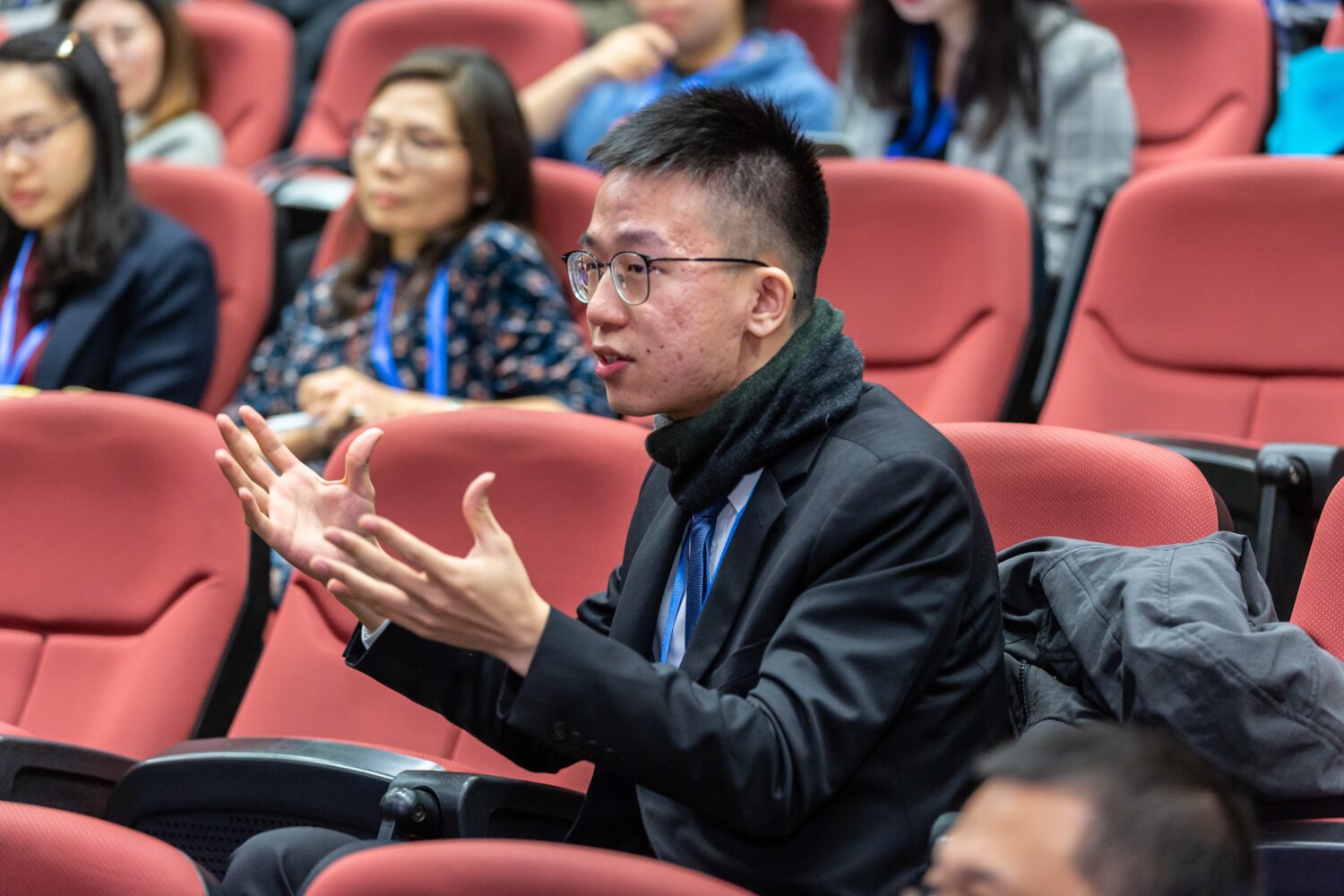 This is a rectangular image. This image appears to be from a presentation hall with some people sitting in the chairs. There is one male person of Asian origin sitting in the audience chair and appears to be taking some hand gestures, other people appear to be listening.
