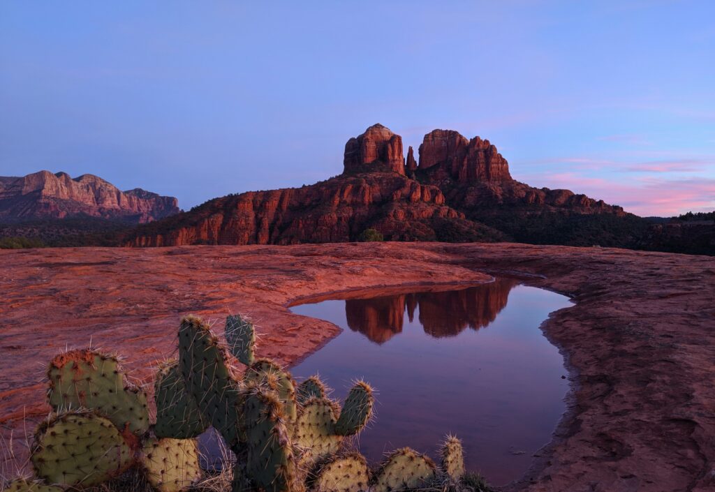 This is a rectangular image capturing the reflection of Cathedral Rock in pools of captured rainwater in the area known as Secret Slick Rock in Sedona, Arizona. The iconic Cathedral Rock formation towers majestically in the background, mirrored perfectly in the still water below. The red rock formations, serene water, and natural beauty of the location combine to create a captivating and tranquil scene.