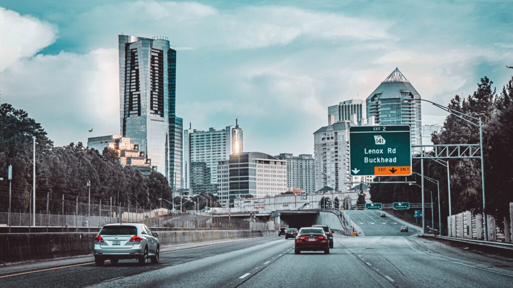 This shows an image taken on a highway that leads into a city with big buildings. This signboard on the highway shows " Lenox Rd, Buckhead"