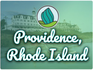 This image shows an empty paddle boat at the shore and in the background, there is a large building structure with a USA flag on top. In the top center, there is the logo of NDC and below there is the text " Providence Rhode Island"
