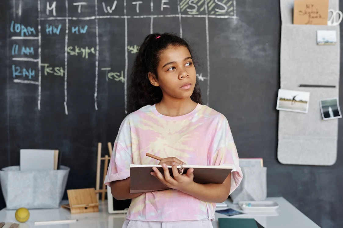 A young person a tie-dye shirt holding a notebook and pencil in front of a blackboard and appears to be in thinking.