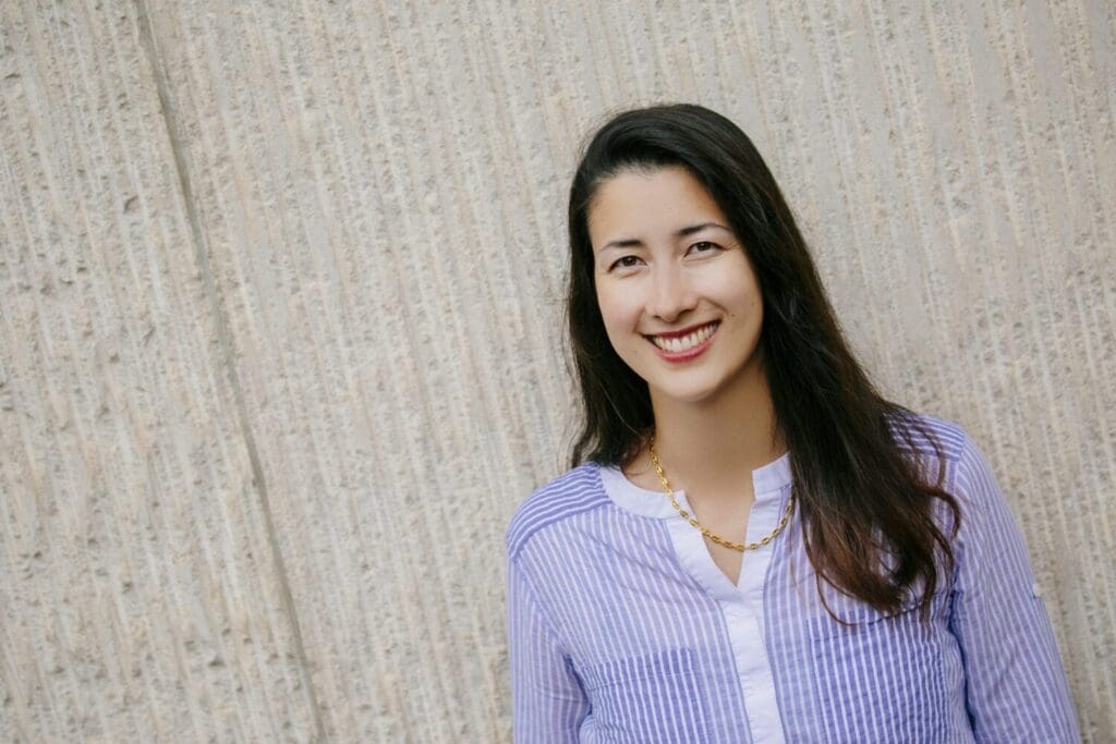 A long brown haired person in blue striped blouse smiling in front of a white wall.