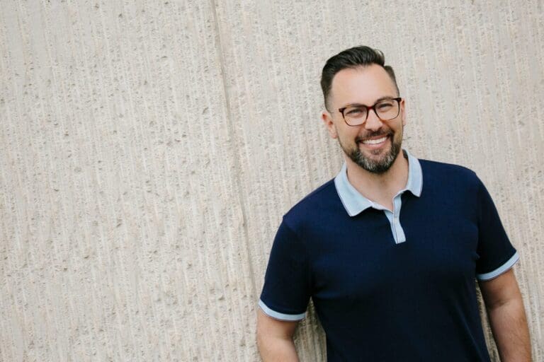 Una persona de cabello corto y castaño, con gafas y camiseta polo azul, sonriendo frente a una pared blanca.