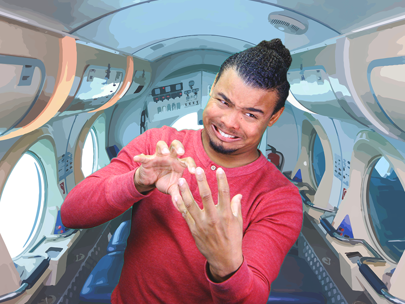 Justin Perez, a black man with long curly black hair pulled back into a bun, a chin goatee, and a red long-sleeved shirt stands in the interior of a spaceship. He faces the camera and is very expressive as he tilts to the right and signs something with two hands facing each other. The interior of the spaceship looks like a long shiny white rounded hallway. There are several large round windows along each wall. One side shows light coming in while the other side is dark. The floor is dark blue with rows of small lights on either side. Near the ceiling are rows of overhead bins with closed doors. The wall behind Justin displays symbols in black, white, and red and there is something that looks like a smoke detector on the ceiling.