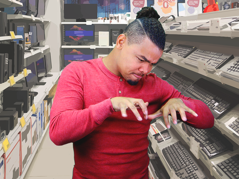 Justin Perez, a black man with long curly black hair pulled back into a bun, a chin goatee, and a red long-sleeved shirt stands in an aisle of an electronics store. Though he faces the camera, he is looking down at his hands as he demonstrates and signs “typing”. On the shelves behind him are rows of black and grey keyboards, computer bases, monitors, white and red boxes, and large yellow sales tags. The shelves and floor are beige. Along the wall behind Justin are more shelves of monitors and computer bases. Just visible near the top are large advertising signs in white, orange, purple, and mauve.