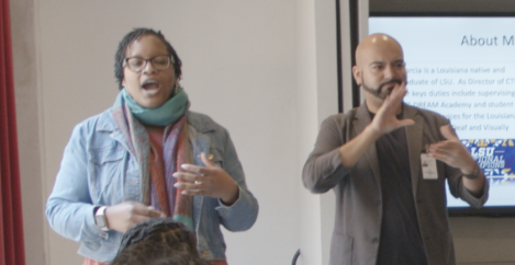The image shows a person in blue jean jacket and glasses speaking and there is another person in a brown jacket interpreting.