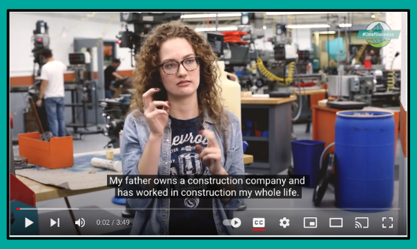 A long curly haired person in glasses talking using sign language and in the background it looks like a mechanical shop full of tools.