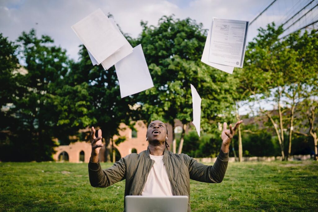 This image shows a student sitting in an open green area with a laptop in front of him & tossing papers in the air.