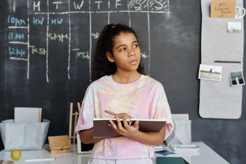 A young person wearing a tie-dye shirt holding a notebook and pencil in front of a blackboard and appears to be in thinking.