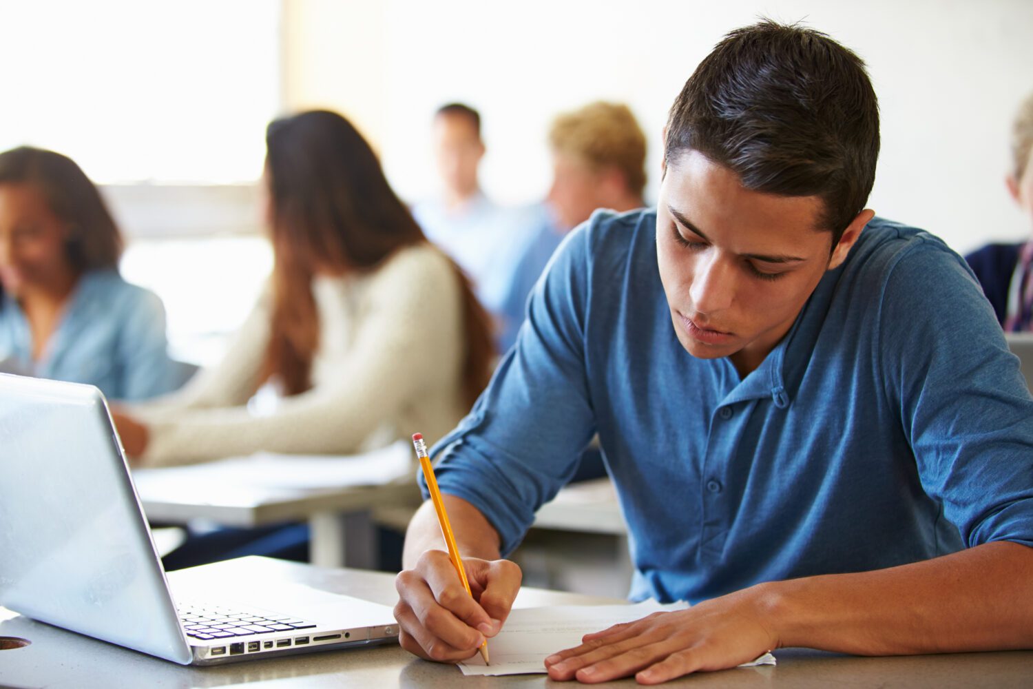 This image appears to be from a classroom. This shows a young male student sitting & appears to be writing on paper with a laptop next to it. He is wearing a blue t-shirt. In the background of the image, there are blurred images of other students.