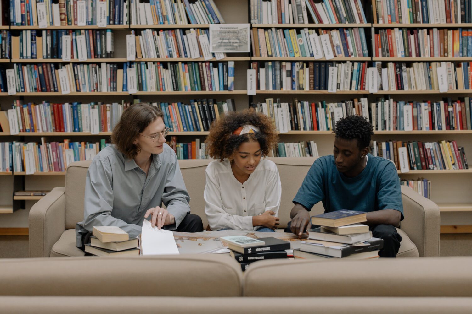 This image is taken in a library. There is a couch and three students sitting on it. There are two male students & one female student. They appear to be discussing something from a book. Behind the couch, there is a large book stand with lots of books.