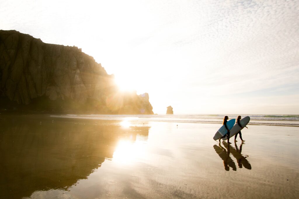 This image is taken at the beach with two people walking with surfing boards in their hands. It's a bright day with the sun coming through a mountain which is on the side of the image.