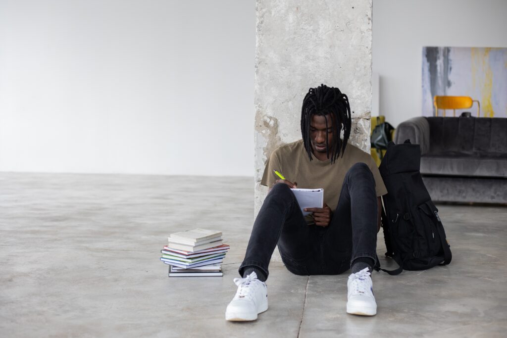 This image shows a male student sitting on the floor while lying on the wall. He appears to be writing something in a book. Next to him, there is a stack of books & a backpack on the other side. He wearing black jeans, white shoes, and a greyish-brown T-shirt.