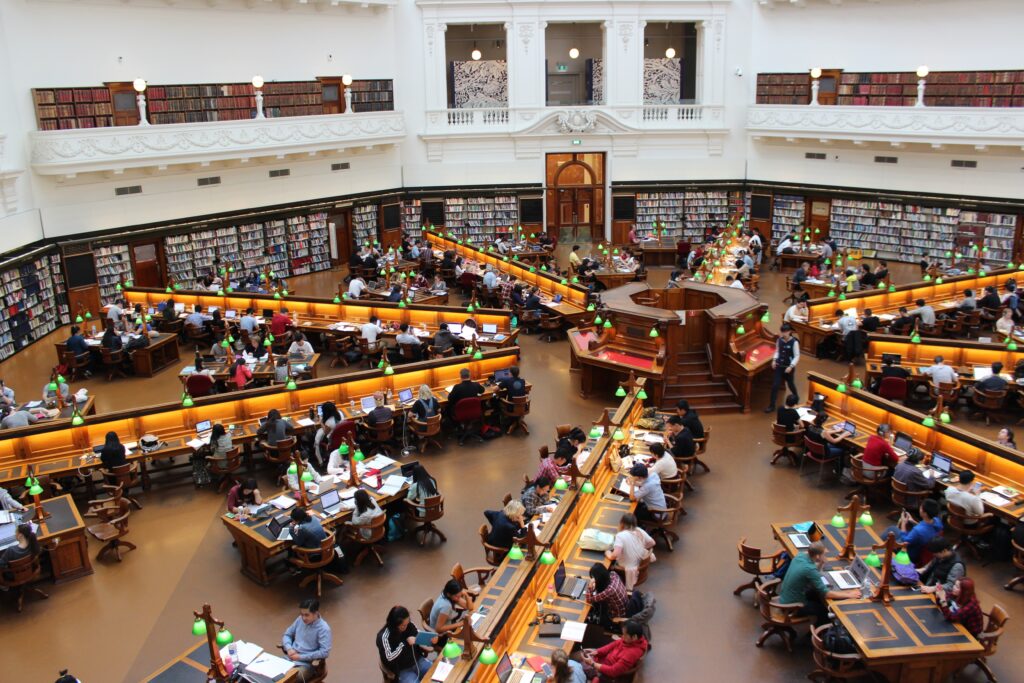 This is a rectangular image showcasing the State Library Victoria. The library is an impressive architectural structure with grand pillars and a dome. In the foreground, a diverse group of people can be seen engaged in various activities, such as reading, studying, and browsing books. The atmosphere is vibrant and intellectual, with individuals from different backgrounds coming together to utilize the resources and spaces provided by the State Library Victoria.