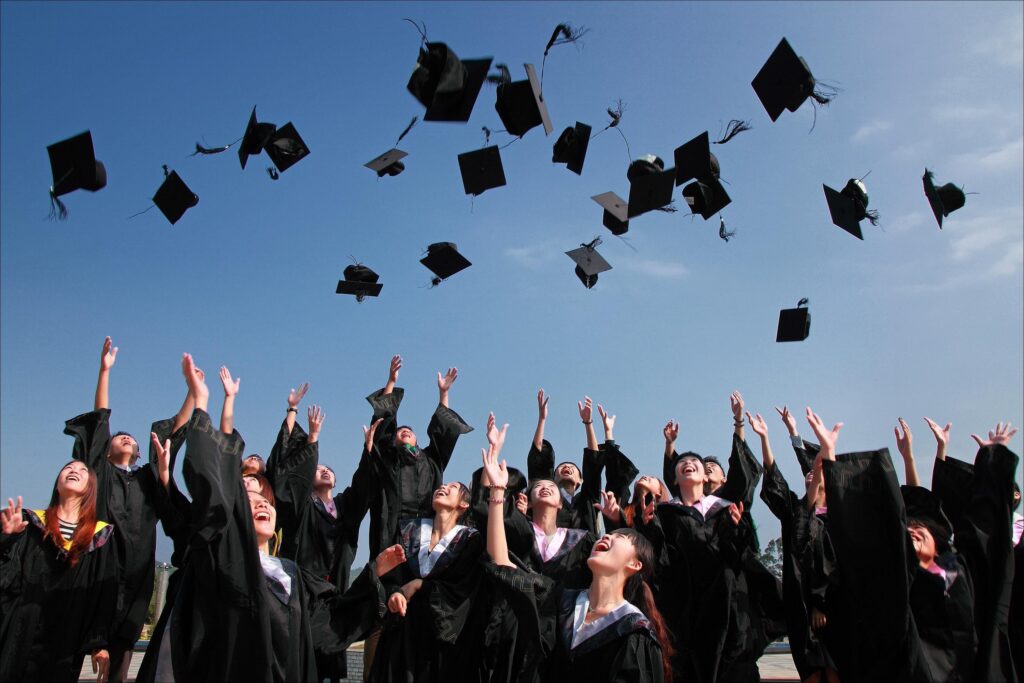 This image is taken in the outside environment. This appears to be a graduation ceremony & students are wearing graduation gowns. They are seen smiling and throwing their graduation cap in the air.