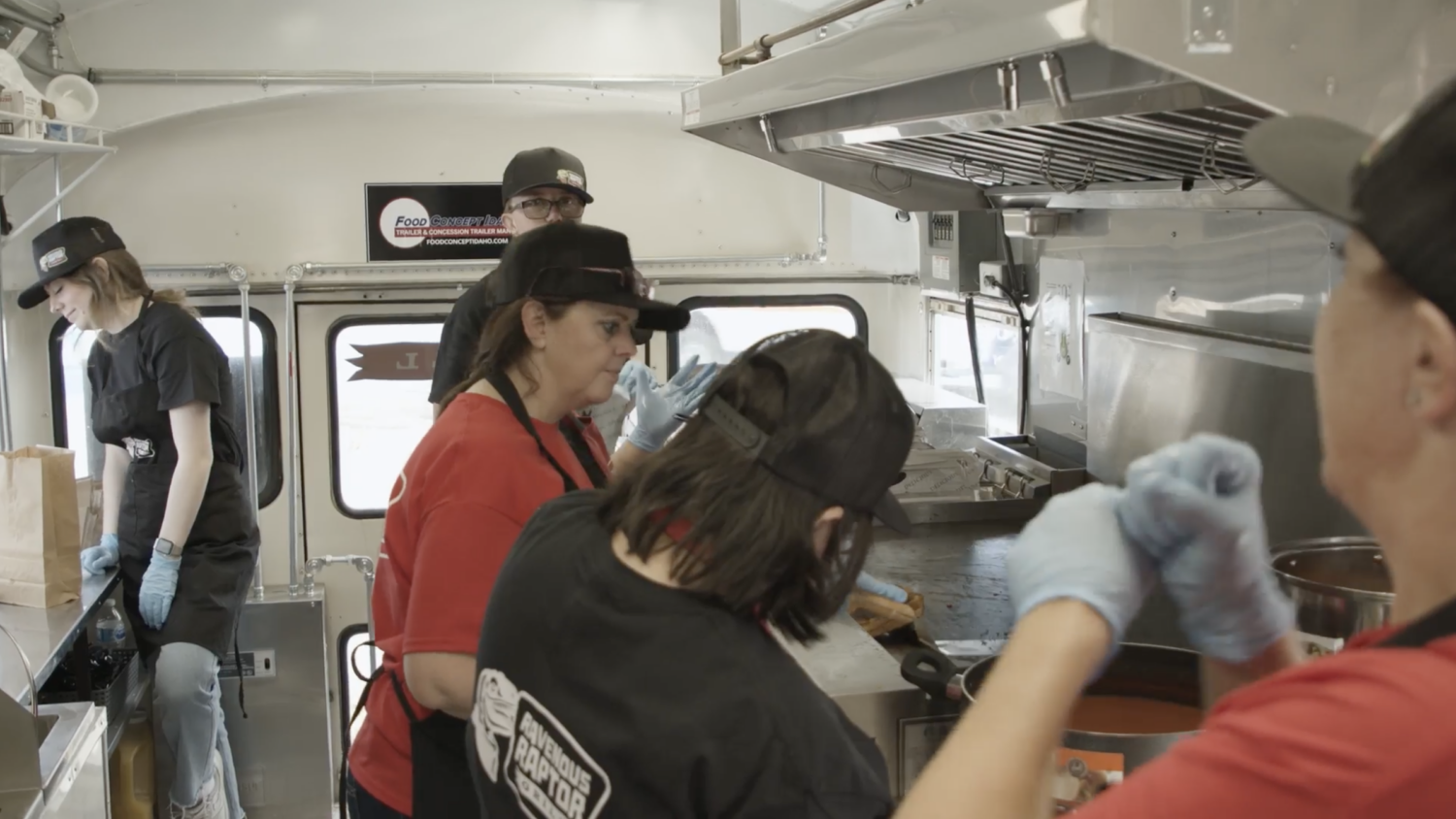 This is a rectangular image of the kitchen area of a food truck. There are five people in the image. Three are wearing black T-shirts & two are wearing reddish color T-shirt. All have a black cap on their heads.