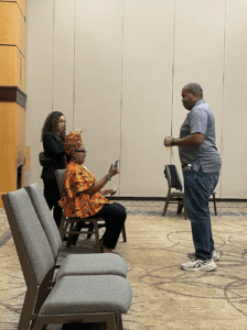 A person sitting on a chair indoors in a colorful shirt and headwrap looking at a phone along with two other person standing up and appears to be having a conversation.