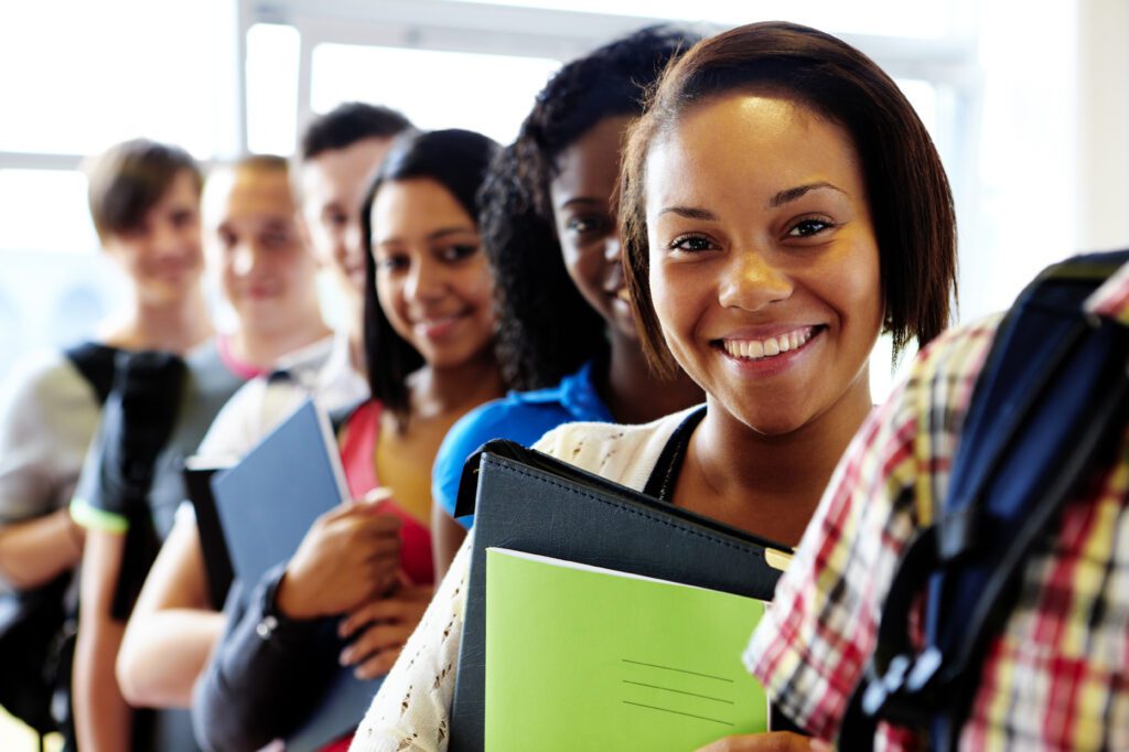 A smiling person with a group of students standing behind her. They are indoors, holding variety notebooks.