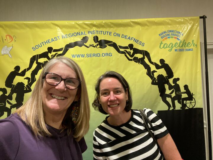 Two people smiling and taking a selfie in front of a banner at the Southeast Regional Institute on Deafness.